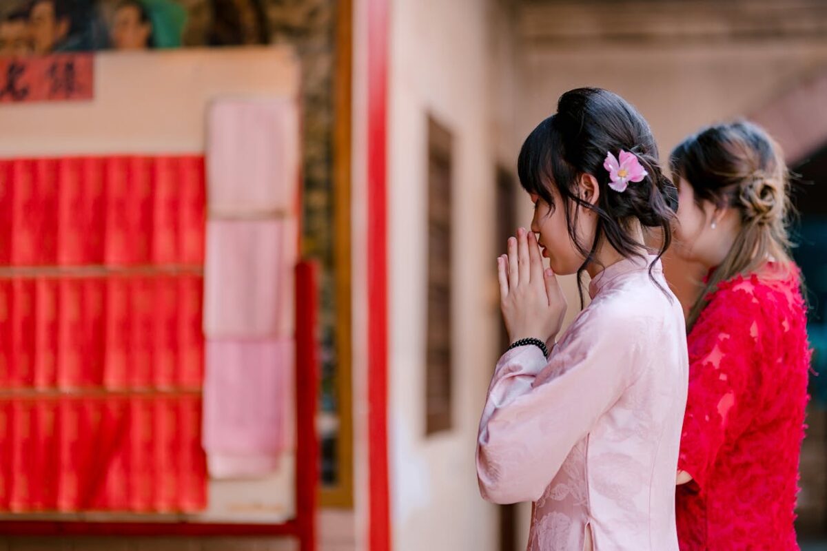 Two women in traditional clothing praying at an indoor Asian temple.