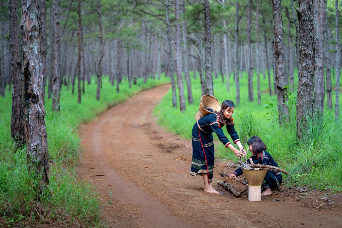 Two ethnic women collecting sticks in a forest path, symbolizing tradition and nature.